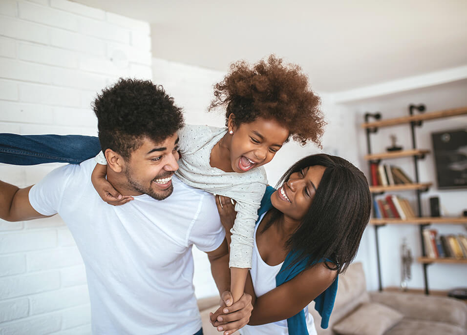 Little girl on her dad's shoulders playing in living room with her parents laughing