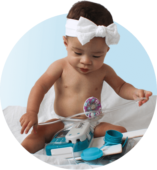Adorable baby girl sitting on floor with her enteral feeding pump in front of her