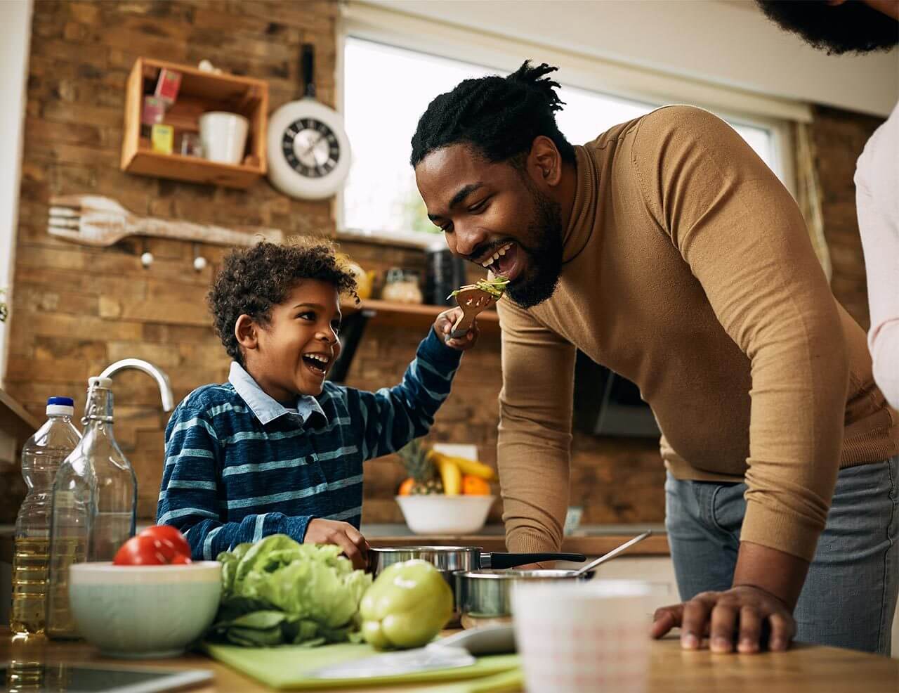 Happy African American boy having fun while feeding his father in the kitchen.