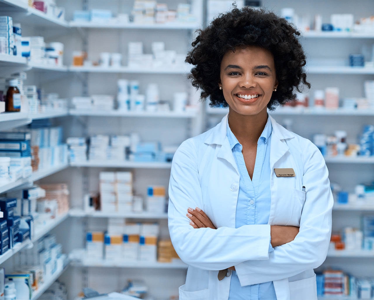 Female pharmacy manager standing smiling with arms folded in front of drug supply closet.